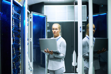 Portrait of young woman working with supercomputer standing in center of server room holding...