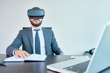 Portrait of handsome modern businessman wearing VR glasses watching 360 videos sitting at desk in office