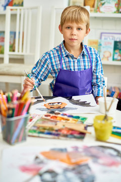 Portrait of blonde little boy looking at camera while painting Halloween pictures sitting at table in art class