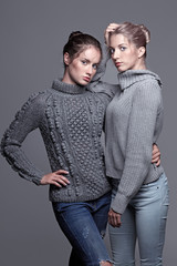 Two young women in gray sweaters on grey background. Beautiful girls stretching hands forward in embrace. Female friendship concept
