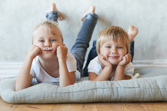 Indoor picture of two blue eyed Caucasian siblings dressed in similar white t-shirts and jeans lying on grey mat on floor barefooted, looking at camera, resting head on their hands and smiling