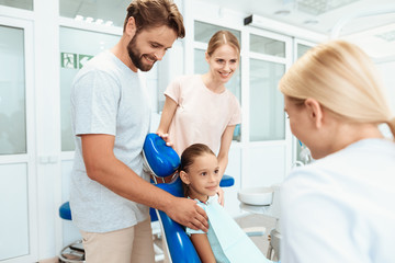 Parents brought their daughter to see a dentist. The girl is sitting in the dental chair. A doctor stands nearby