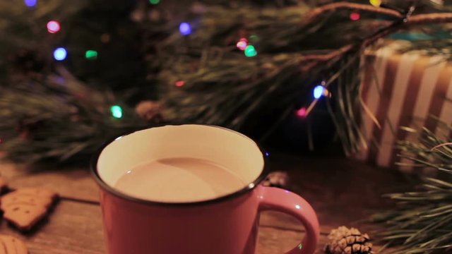 Delicious Christmas holiday with latte. Just stired up hot drink stays on wooden table near decoration from illuminated pine, gift box and gingerbread scones nearby, close up