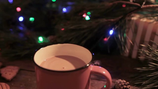 Delicious Christmas holiday with cup of latte. Just stired up hot drink stays on wooden table on festive background of illuminated pine, gift box and gingerbread scones nearby, close up