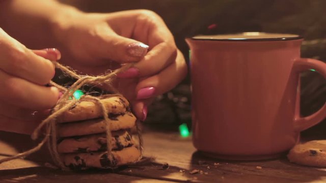 Delicious Christmas holiday with latte and cookies. Close up woman binding sweet chocolate scones near cup of hot drink on illuminate fairy lights background. Cozy xmas evening concept