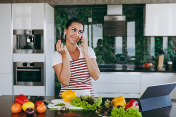 Young happy woman in apron talking on mobile phone in kitchen with laptop on the table. Vegetable salad. Diet. Dieting concept. Healthy lifestyle. Cooking at home. Prepare food.