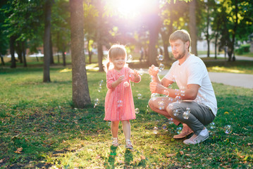 happy father with a small daughter at sunset blowing soap bubbles in a summer park outdoors