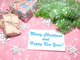 Christmas greeting card over light pink table with snowflakes, gifts, fir tree branches. Flat lay style