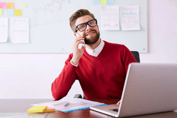 Young businessman talking on phone while in office working with laptop