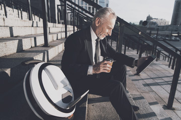 A respectable old man in a strict business suit sits on steps of office. He works on a tablet, next to his mono-wheel © Vadym Huzhva