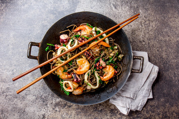 Buckwheat stir-fry noodles with seafood - shrimps, octopus, squid in cast iron asian wok with cooking chopstick. Top view, stone background