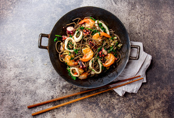 Buckwheat stir-fry noodles with seafood - shrimps, octopus, squid in cast iron asian wok with cooking chopstick. Top view, stone background