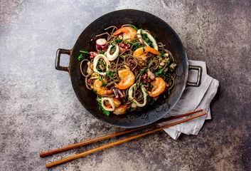 Foto op Canvas Buckwheat stir-fry noodles with seafood - shrimps, octopus, squid in cast iron asian wok with cooking chopstick. Top view, stone background © lblinova