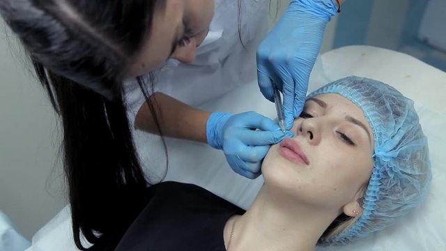The doctor-cosmetologist makes the young girl lip enlargement procedures. Botox injection in the clinic.