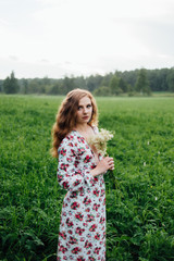 A beautiful girl in a colorful dress stands in the evening on a meadow against a background of fog.