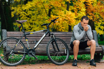 Young pretty athletic man sitting on bench near bicycle in colorful autumn park. Fall season background. Male cyclist resting after ride. Man warming hands with his breath