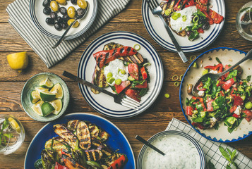 Flat-lay of healthy dinner table setting. Fresh salad, grilled vegetables with yogurt sauce, pickled olives, lemon water over wooden background, top view. Clean eating, vegan, vegetarian food concept