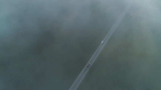 AERIAL, TOP DOWN: White semi truck and delivery van driving along empty country highway through agricultural meadow fields on scary foggy winter morning. Vehicles shipping goods on bad weather day