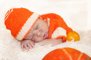 Newborn baby in knitted orange costume sleeping on white blanket with pumpkin and oranges in front of him. Halloween or Thanksgiving concept.