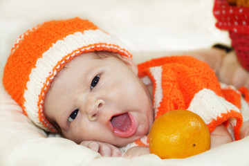 Cute yawning newborn baby dressed in a knitted orange costume with orange in in front of  him. Autumn halloween or harvest concept. Baby face expression.