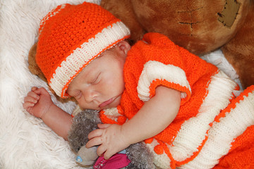 Portrait of cute newborn baby in orange costume sleeping on white fur blanket hugging plush bear. 