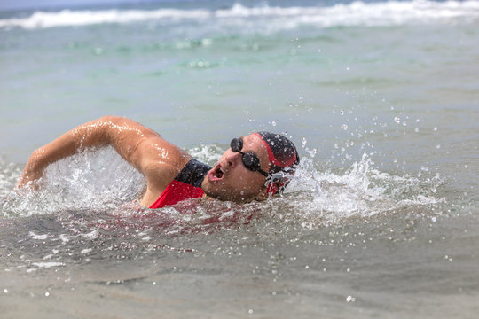 Professional triathlete man swimming crawl ocean freestyle crawl in ocean. Male triathlon swimmer wearing cap, goggles and red triathlon tri suit training for ironman breathing out of water.