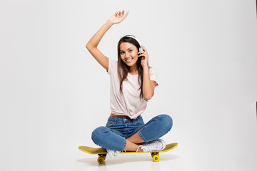 Photo of young smiling brunette woman in white headphones with arm extended upwards, looking at camera while sitting on yellow skateboard