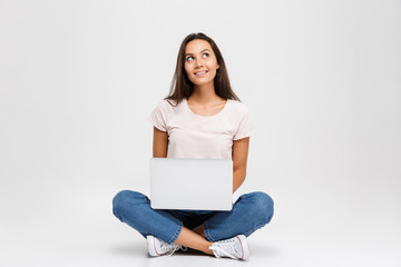 Photo of young thinking brunette woman, holding and using laptop, while sitting with crossed legs