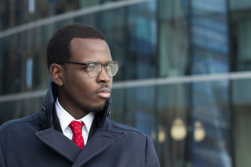 Fototapeta premium Outdoor portrait of handsome African businessman pictured in urban environment dressed in coat with lifted collar looking to street through eyeglasses with expression of serious concentration on work