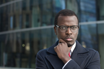 Urban portrait of handsome African businessman dressed in smart coat in city street pressing fist to his chin while thinking hard and trying to make best decision for development of his business