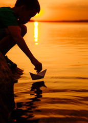 Little boy launch paper ship on the water. Beautiful summer sunset. Paper boat. Origami.