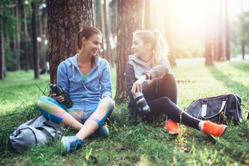 Two young women in sportswear sitting under trees in the forest drinking water, talking and resting after training outdoors