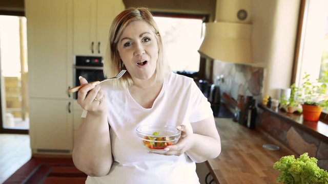 Overweight woman at home eating vegetable salad in the kitchen.