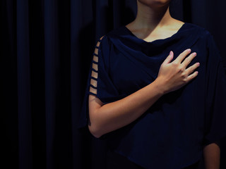 Woman wearing blue black shirt with shoulder hole details, with black curtain background, with one right hand putting on her chest, as to feel her heart pain or deeply sad in love