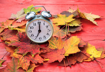 metal clock standing on a red wooden table amongst fallen autumn leaves a bright Sunny autumn morning