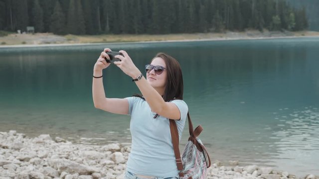 Young Woman Taking Selfie By Smartphone In Front of Mountain Lake. Beautiful Caucasian Girl Spending Time In a Moutain Forest