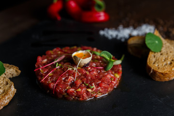 Beef tartar with a quail egg on top, grilled bread, greenery, red chilly pepper, salt and spices, served on a black stone plate.