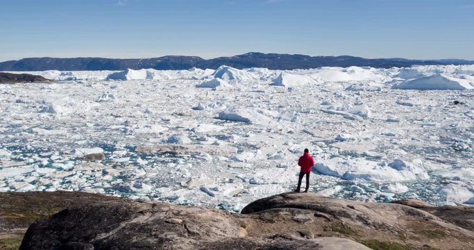 Travel wanderlust adventure in Arctic landscape nature with icebergs - tourist person looking at view of Greenland icefjord - aerial video. Man by ice and iceberg, Ilulissat Icefjord.