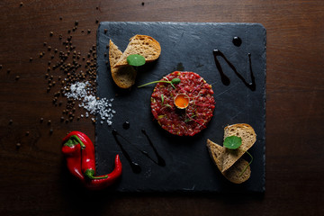 Beef tartar with a quail egg on top, grilled bread, greenery, red chilly pepper, salt and spices, served on a black stone plate.