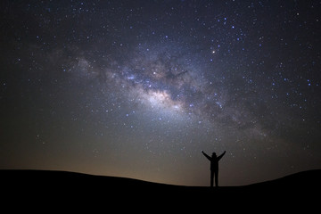 Fototapeta premium Landscape with milky way galaxy, Starry night sky with stars and silhouette of a standing sporty man with raised up arms on high mountain.