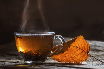 Transparent cup with tea and autumn yellow dry leaf on a dark background. The smoke is curly from the cup. Autumn mood of coziness and warm atmosphere.
