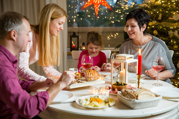 Happy family celebrating holiday together, sitting around decorated round table. Mother serving dinner. Living room with Christmas tree, lights and candles. Merry Christmas concept
