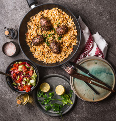 Meat balls with vegetables rice in pan and salad, served on gray stone kitchen table background with plates and cutlery, top view