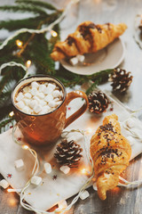 hot cocoa with marshmallows and croissant on rustic wooden table with christmas lights. Cozy winter home concept