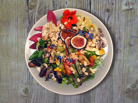 Üppiger bunter Salat mit vielen Früchten und essbaren Blüten in Steingutteller auf altem Holztisch