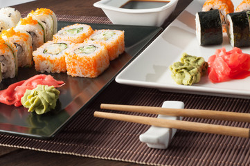 Sushi rolls on a black and white plate, on a wooden mat with chopsticks and soy sauce