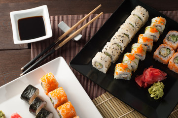 Sushi rolls on a black and white plate, on a wooden mat with chopsticks and soy sauce