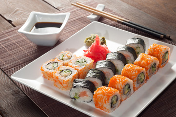 Sushi rolls on a black and white plate, on a wooden mat with chopsticks and soy sauce
