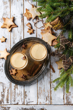 Eggnog Christmas milk cocktail with cinnamon, served in two glasses with shortbread star shape sugar cookies different size, decor toys, fir branch over white wooden plank table. Top view with space