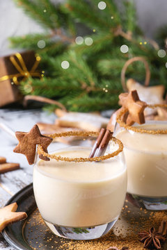 Eggnog Christmas milk cocktail with cinnamon, served in two glasses on vintage tray with shortbread star shape sugar cookies, decor toys, fir branch over white wooden plank table. Close up